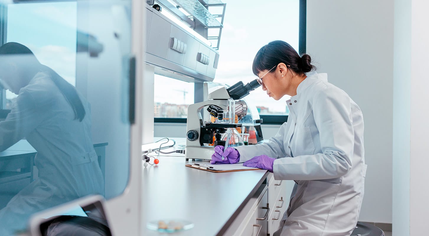 A woman wearing a lab coat inspects a specimen under the microscope, surrounded by laboratory equipment.